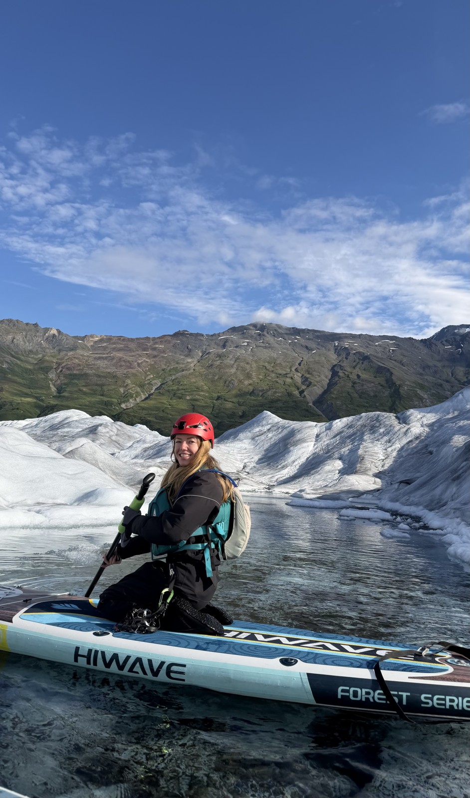Anya paddleboarding beside a glacier, wearing a red helmet