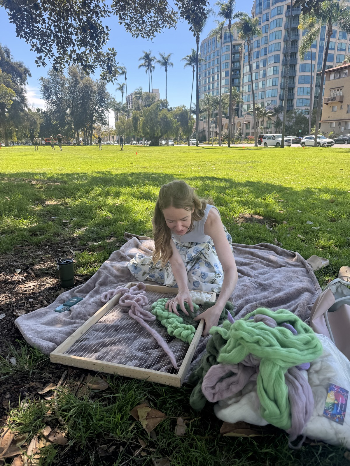 Anya weaving with colorful wool on a picnic blanket in a park
